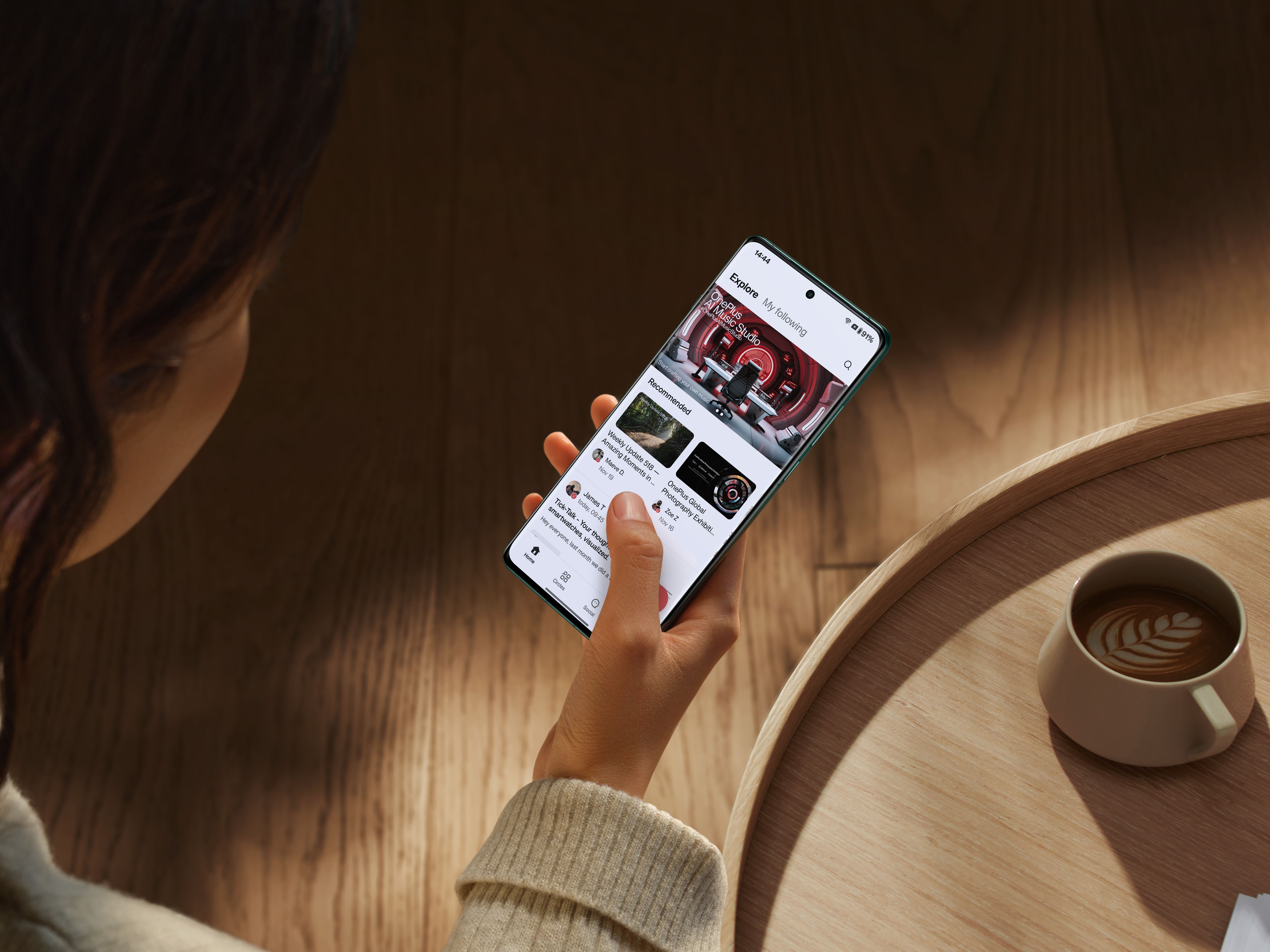 Человек держит OnePlus 12 в одной руке над деревянным столом, подносом и чашкой кофе.